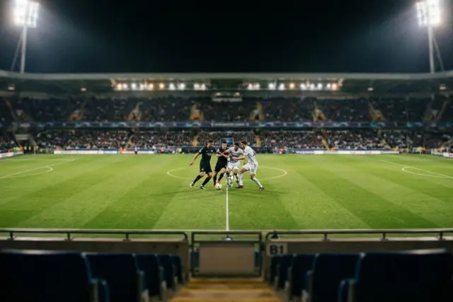 Wide view from stadium seats of a night soccer game. Four players contest the ball at midfield under bright